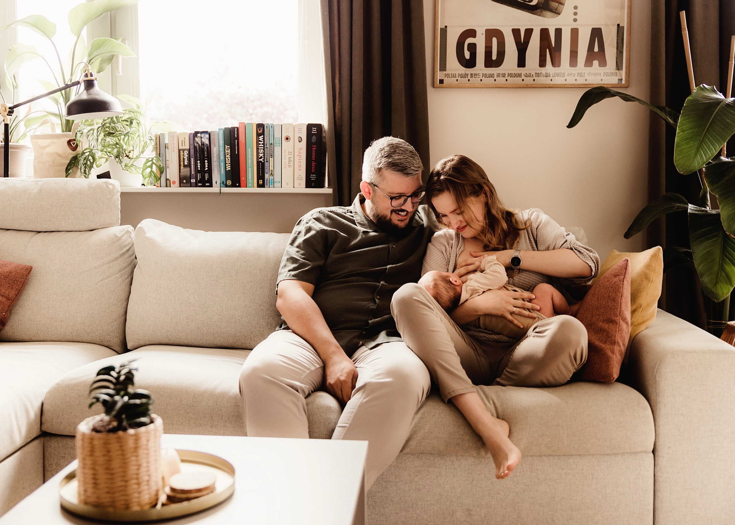 Domowa sesja noworodkowa lifestyle, Agnieszka i Krzysiek z małym Jerzykiem na kanapie, mama karmiąca niemowlę piersią w przytulnym salonie, naturalna fotografia rodzinna Gdynia