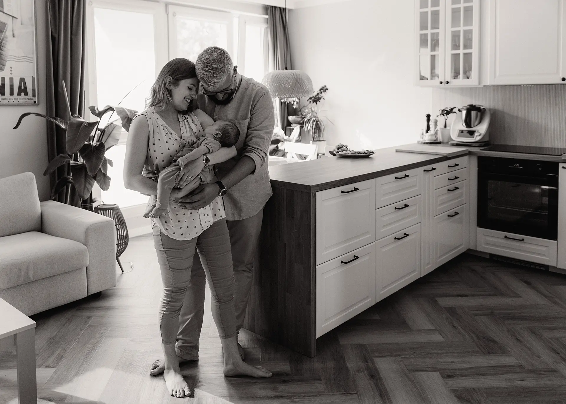 Czarno-biała fotografia lifestyle z domowej sesji noworodkowej w Sopocie. Agnieszka i Krzysiek trzymają małego Jerzyka w nowoczesnej kuchni połączonej z salonem. Naturalne światło i autentyczne emocje.