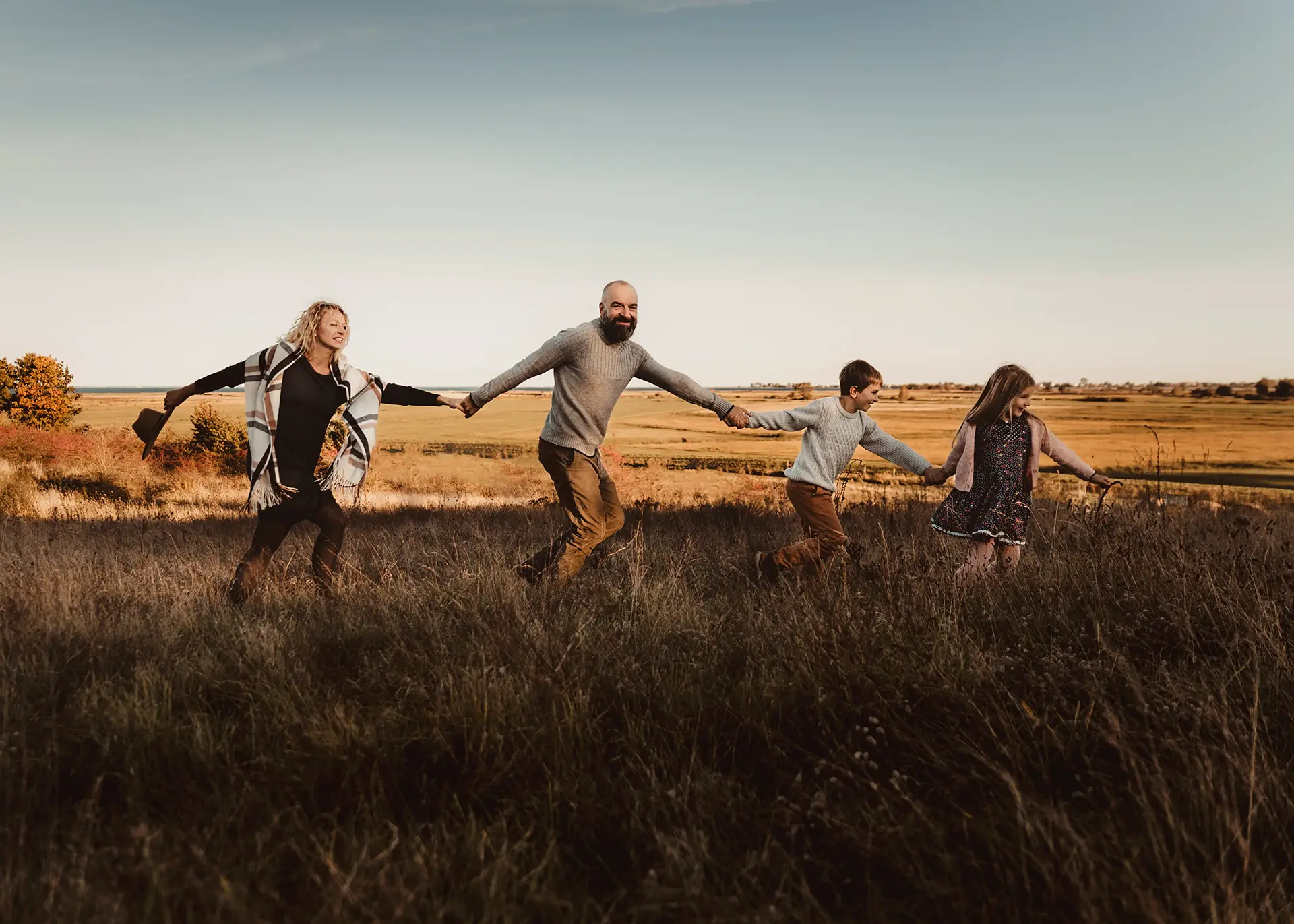 Radosna sesja rodzinna w plenerze o zachodzie słońca, rodzice i dwoje dzieci biegnący za ręce po łące, naturalna fotografia lifestyle Gdańsk