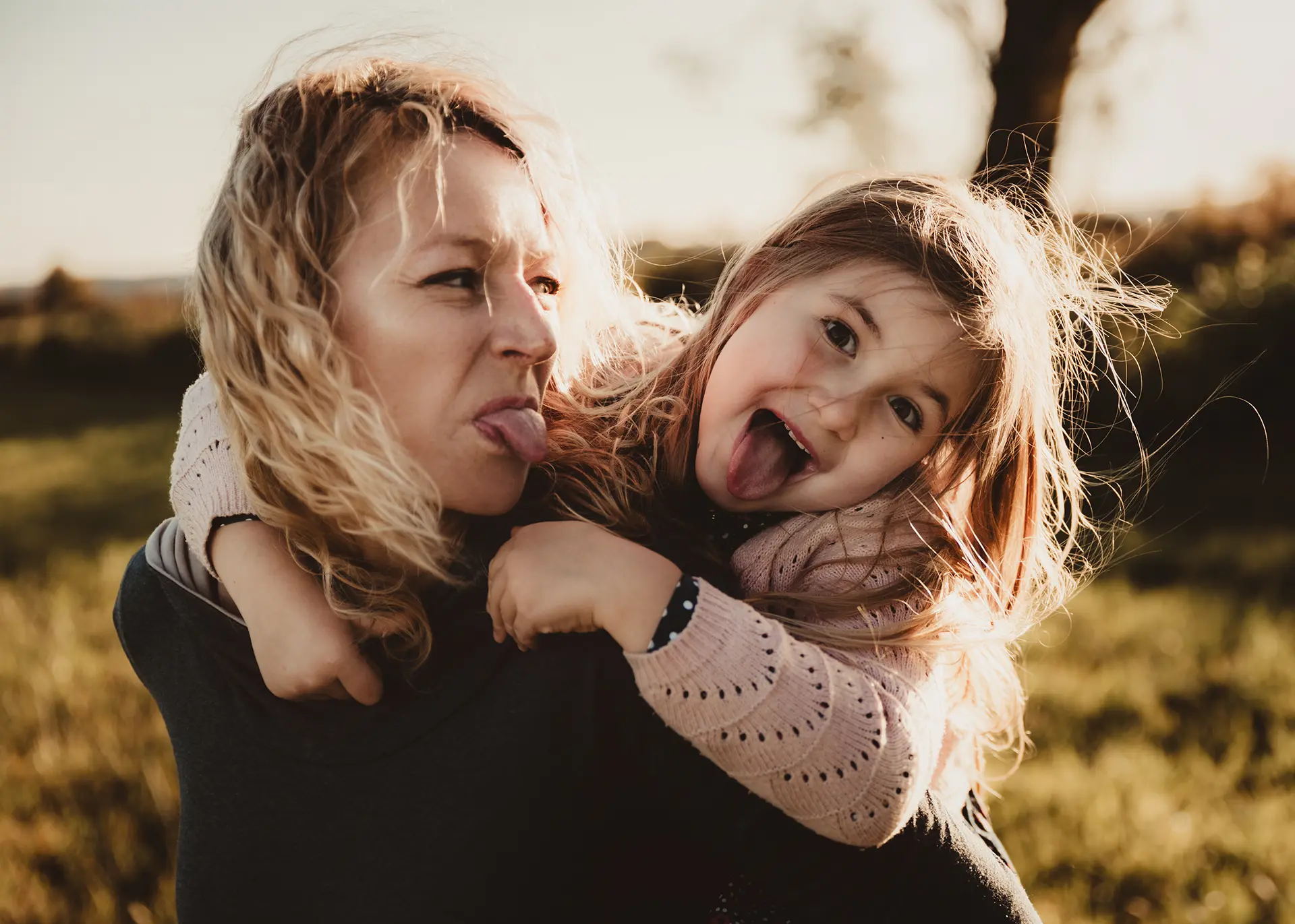 Bliski portret mamy i córki strojących miny z wystawionymi językami podczas radosnej sesji rodzinnej w plenerze, złota godzina i naturalne emocje, fotograf rodzinny Gdańsk