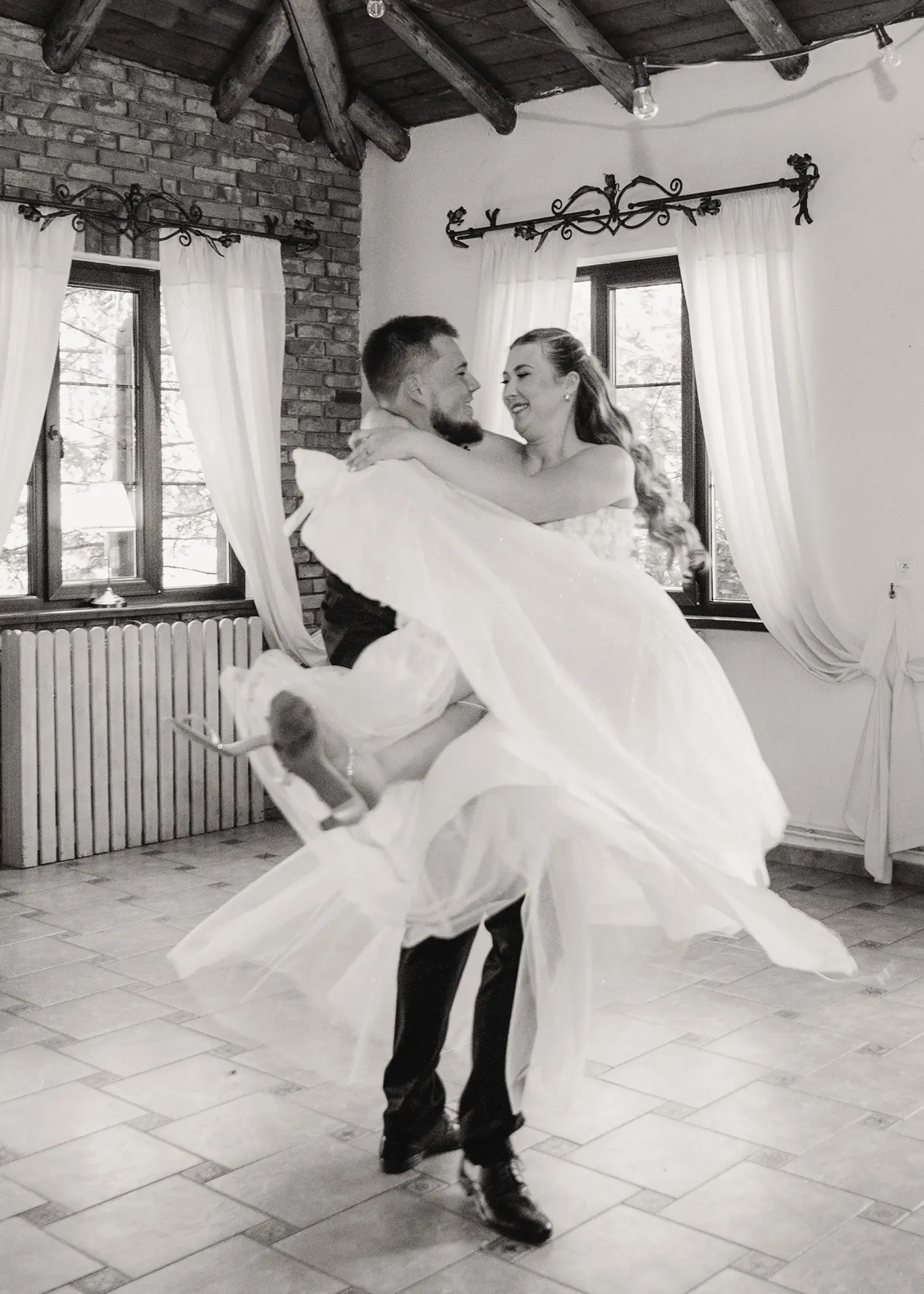 Czarno-biała fotografia ślubna, pan młody biorący pannę młodą na ręce podczas pierwszego tańca, roześmiana para młoda na sali weselnej, dynamiczny reportaż ślubny Gdynia