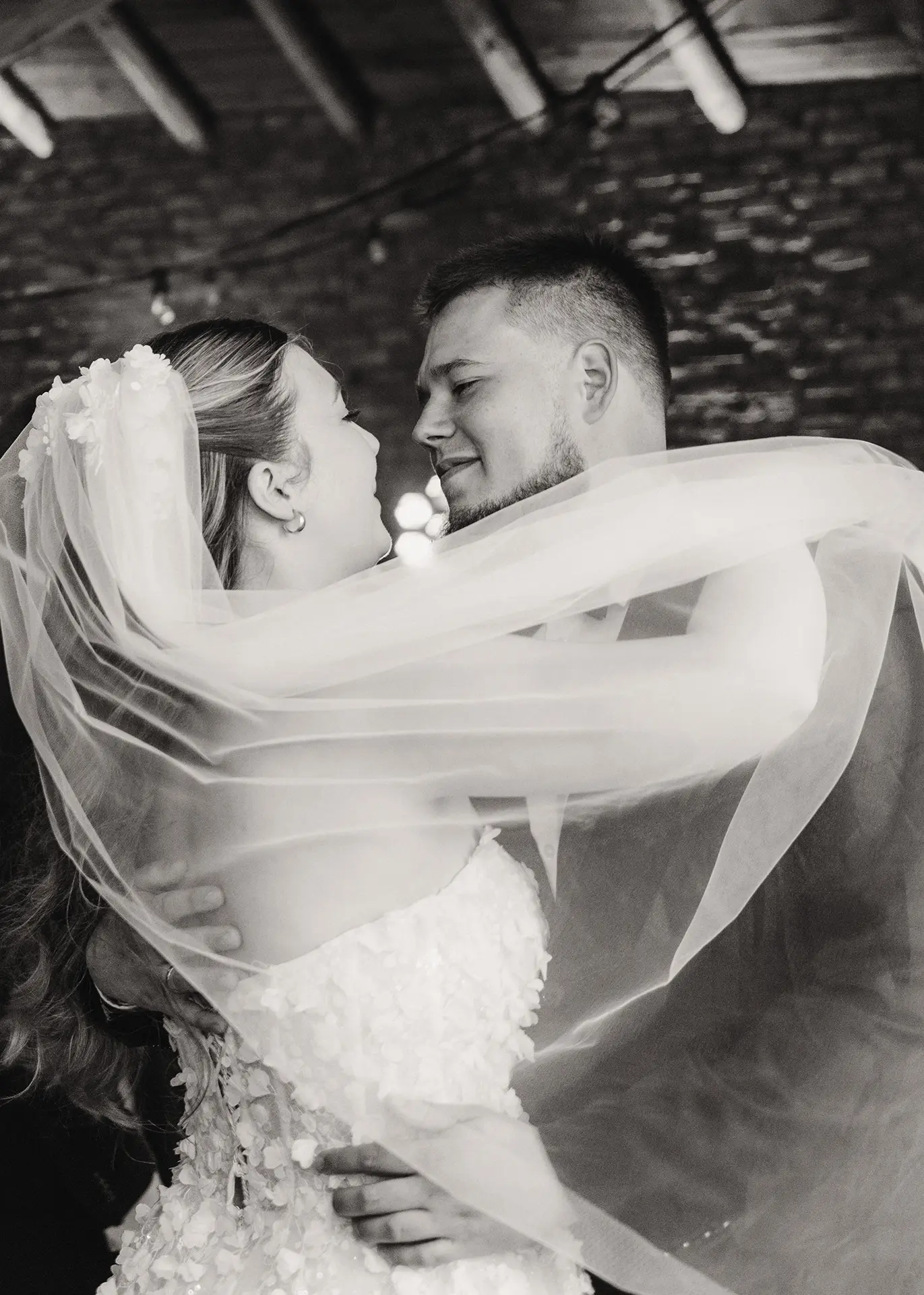 Czarno-biała fotografia reportażowa z pierwszego tańca pary młodej. Uśmiechnięty pan młody trzyma pannę młodą, która jest owinięta welonem. Dynamiczne ujęcie, zabawa weselna Gdańsk.